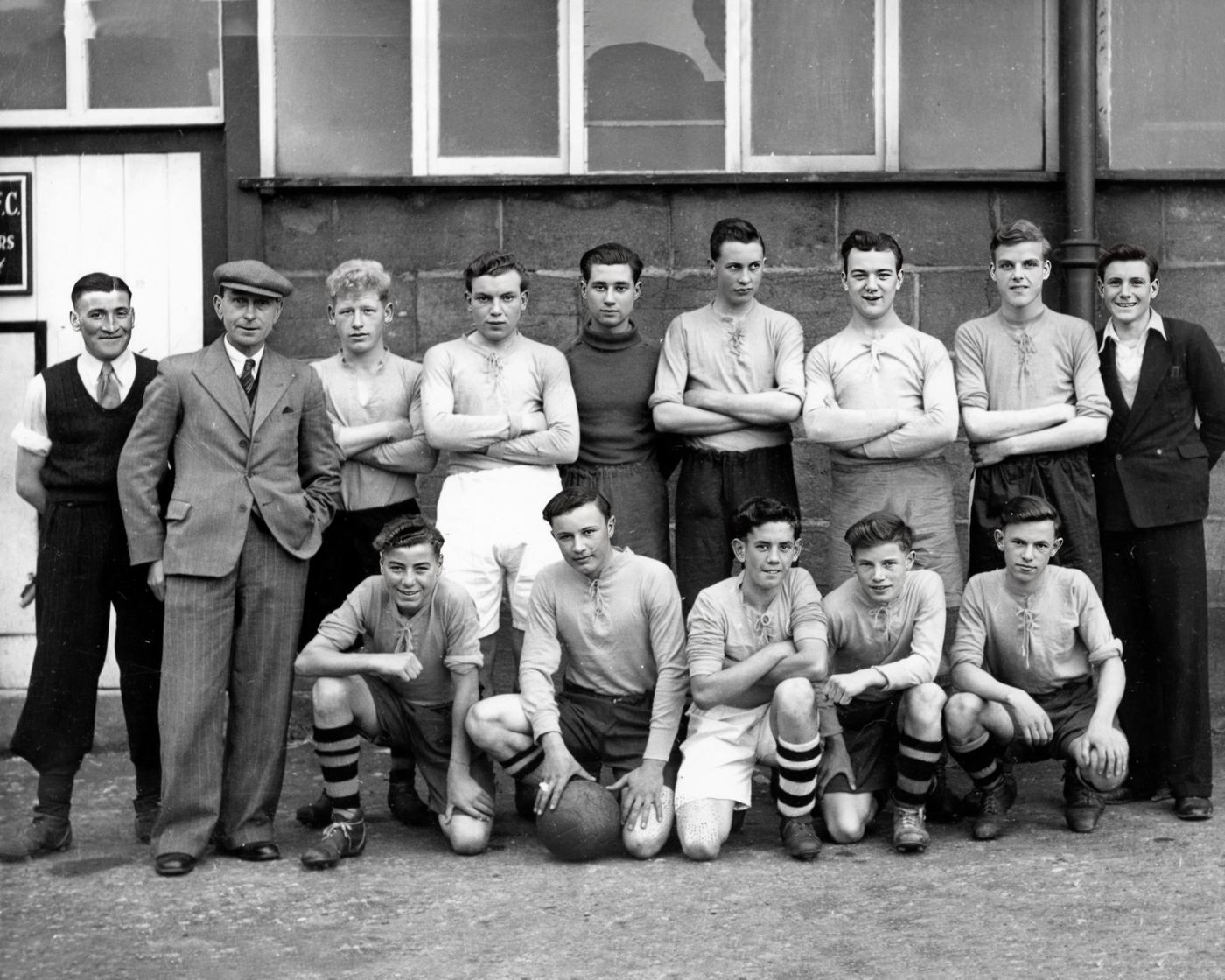 Young players in light shirts lined up against a stone wall with two men in suits and caps