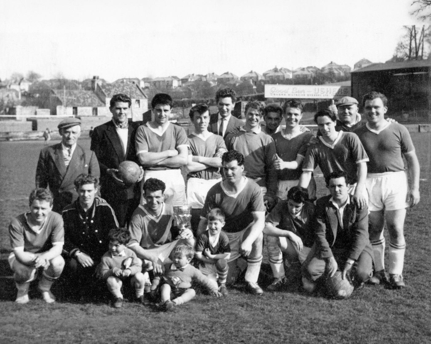Players in dark shirts with a small cup and children in front, with a Good Beer Ushers sign behind
