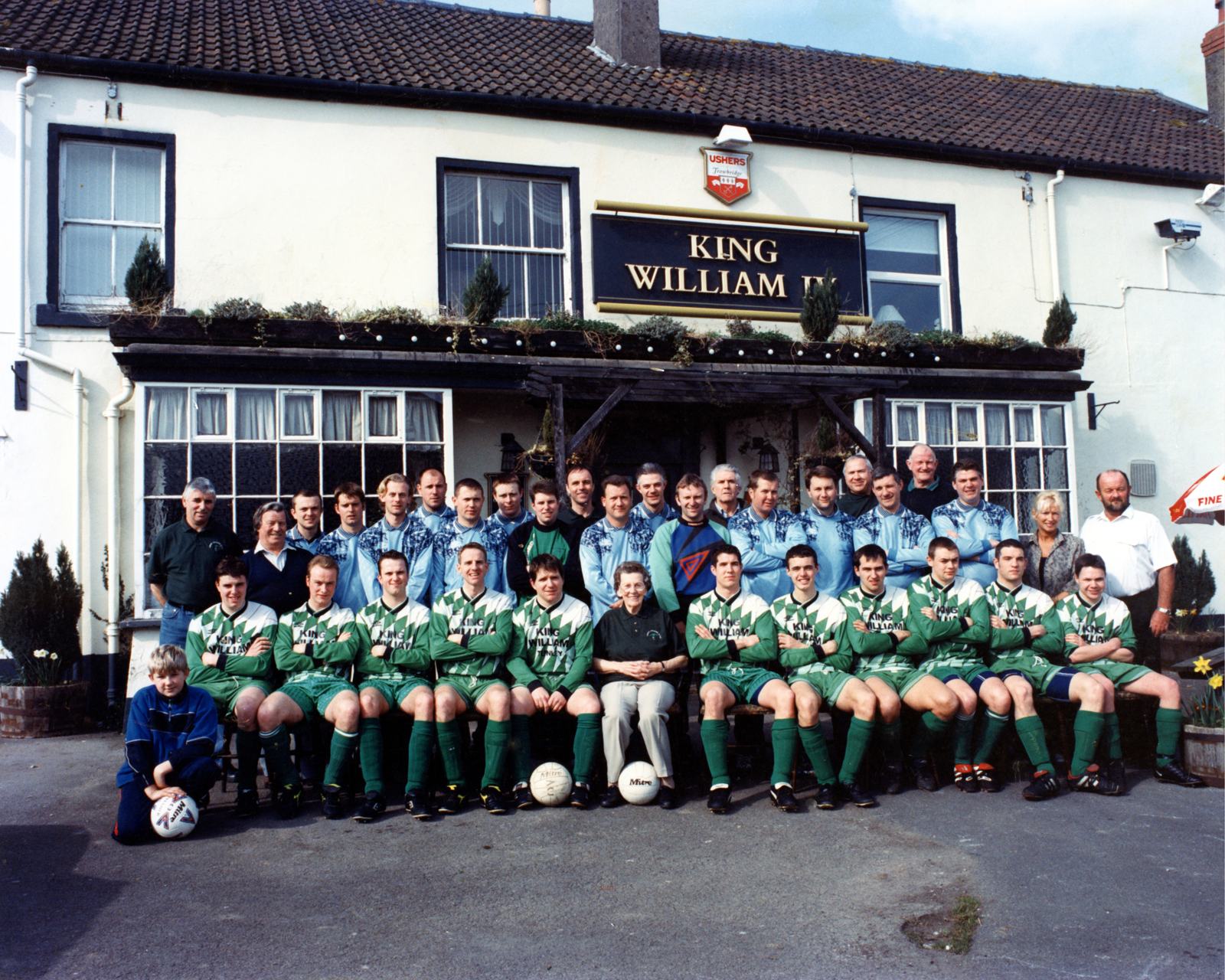 Two Tunley teams in green kit and light blue tracksuit tops, seated and standing outside the King William IV, with an older woman seated in the middle