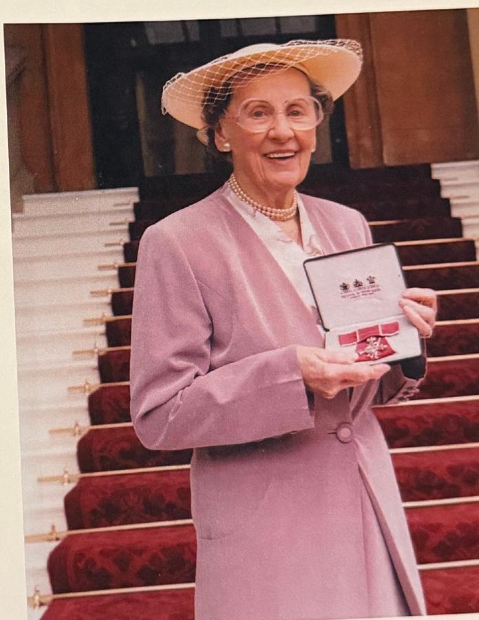 Freda Hookings in a pink coat and hat on red-carpeted steps, holding her MBE in its case