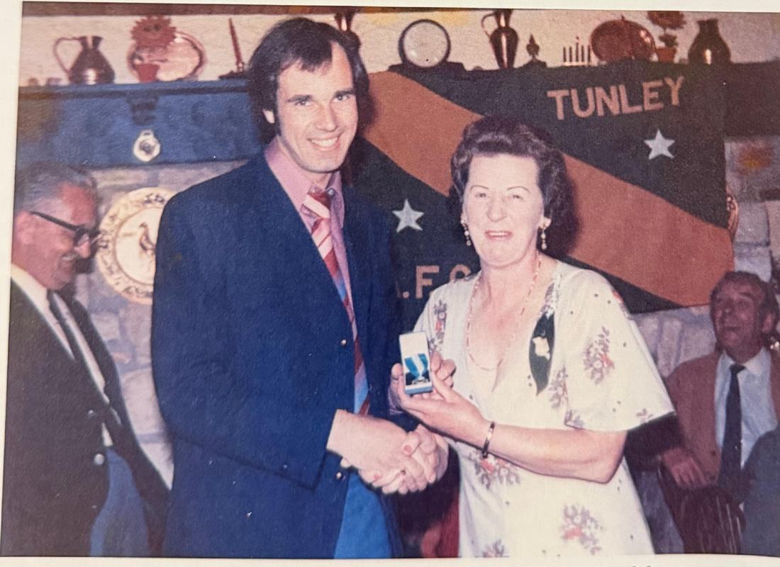 Freda Hookings shaking hands with Alan Pickering as she presents an award, with a Tunley banner behind